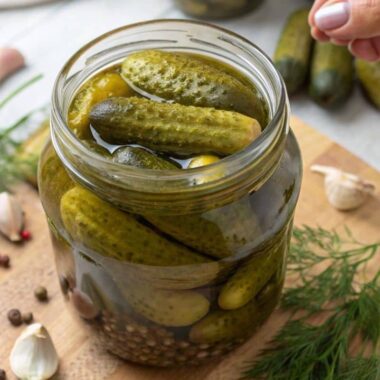 A jar of pickled baby cucumbers surrounded by garlic, dill, and mustard seeds.