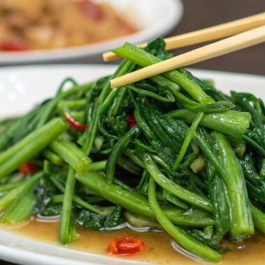 A plate of stir-fried water spinach with garlic, glistening in a light sauce, served on a white plate with a hand holding chopsticks.