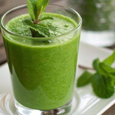 A glass of green smoothie made with raw water spinach, garnished with a sprig of mint and served on a white plate.