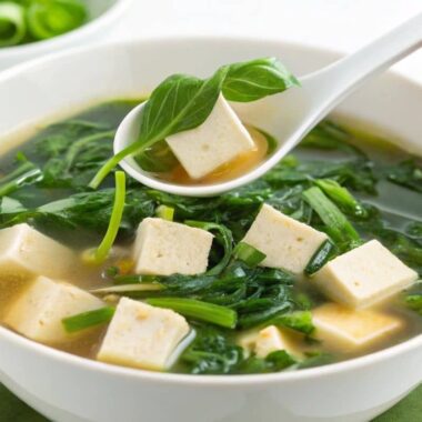 A bowl of steaming water spinach and tofu soup with a spoon lifting tofu and greens