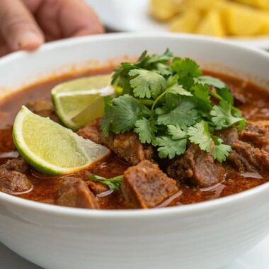 Close-up of a bowl of menudo with beef tripe, chili broth, and garnishes, held by hand