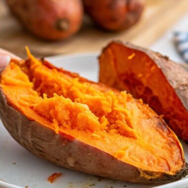 Close-up of a roasted sweet potato on a white plate with a hand holding a slice.