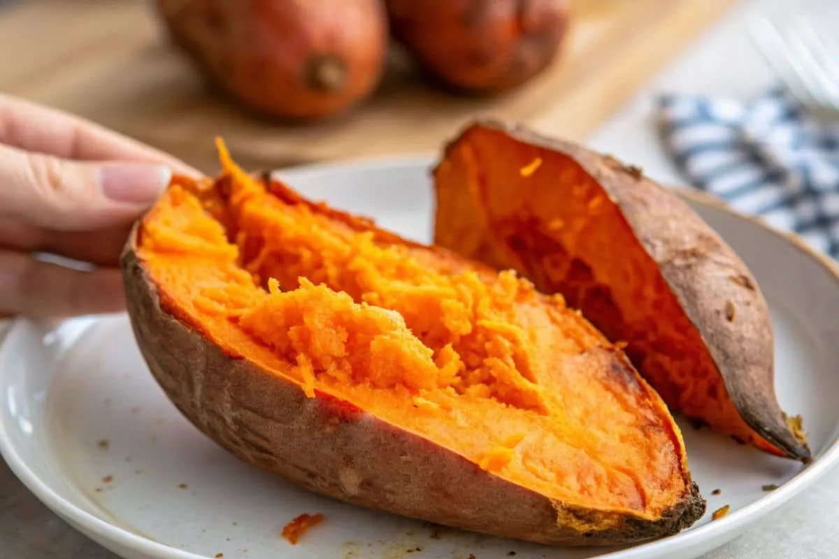 Close-up of a roasted sweet potato on a white plate with a hand holding a slice.